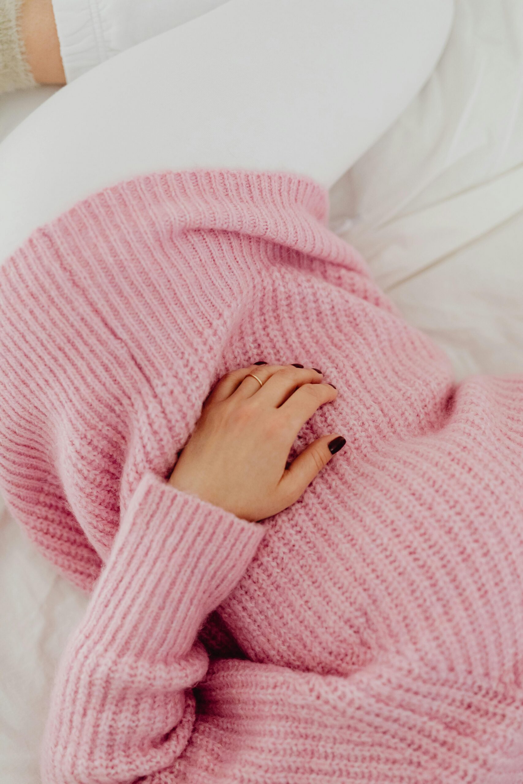 Person in a pink sweater lying down on a white bed with a comfortable and cozy vibe.